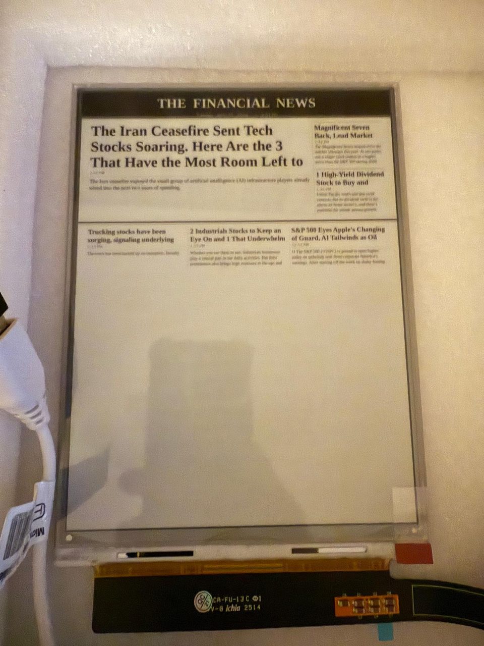 A bare 7.8-inch e-ink panel taped to the wall, showing a freshly rendered front page; the ribbon cable and driver board hang below it.